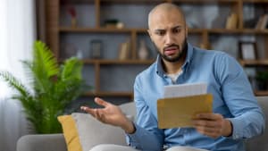Un homme assis sur un canapé à la maison, ouvrant une lettre reçue et ayant l'air préoccupé.
