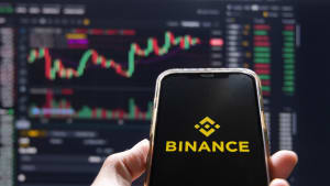 Vue d?une main tenant un téléphone portable avec le logo de Binance à l?écran, sur fond de graphique du Bitcoin