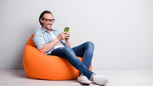 Un homme souriant consulte son smartphone assis sur un pouf