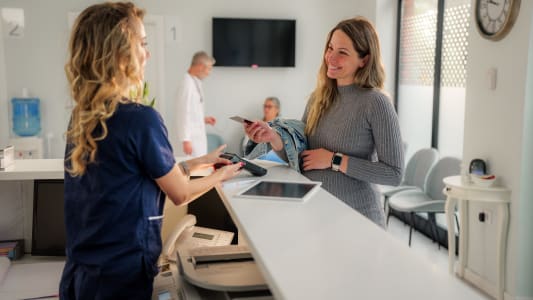 Une femme enceinte effectue une transaction de paiement sans contact au comptoir d'accueil d'une clinique mdicale moderne, interagissant avec la rceptionniste pendant qu'un mdecin se promne en arrire-plan