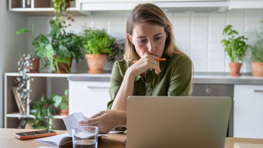 Une femme pensive examine les factures avec un ordinateur portable et un verre d'eau dans la cuisine de sa maison.