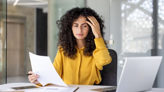 Une femme préoccupée examine des documents dans son bureau, apparaissant stressée pendant qu'elle travaille sur son ordinateur portable et analyse des papiers sur son bureau.