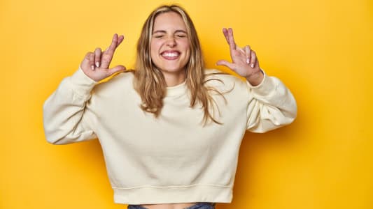 Jeune femme blonde en sweatshirt blanc sur un fond de studio jaune, croisant les doigts pour avoir de la chance