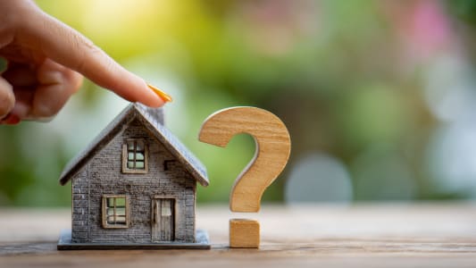 Doigt pointant une maison miniature et un point d'interrogation en bois.