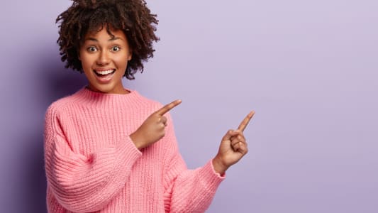 Plan d'une femme charmante et positive, qui montre quelque chose dans un espace vide, a une expression de joie, porte un pull rose ample, fait des gestes sur un mur violet.