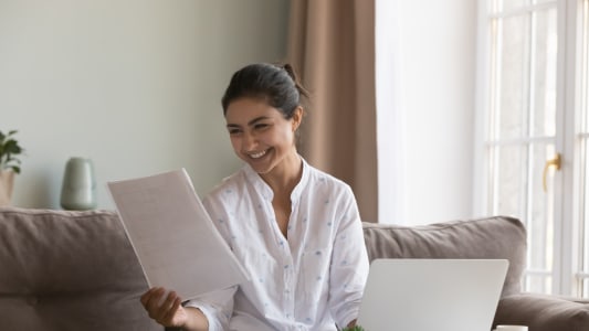 Une femme souriante regarde un document devant son ordinateur porbable.