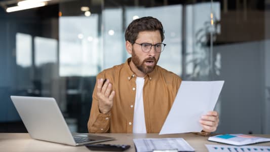 Un hommr assis � son bureau, tenant des documents et r�agissant avec une expression choqu�e.