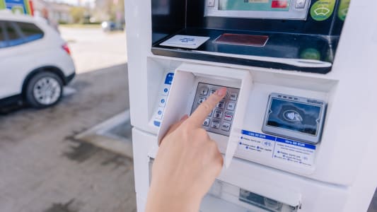 Une personne saisissant un code PIN sur un clavier à un terminal de paiement de station-service tout en ravitaillant une voiture.