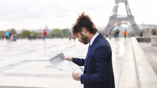 Homme en costume lit un journal devant la tour Eiffel.