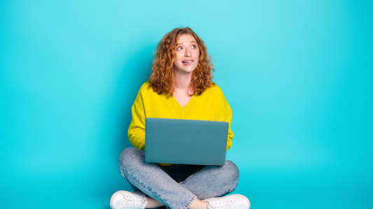 Jeune femme aux cheveux rouges assise en tailleur utilisant un ordinateur portable sur un fond turquoise vif, pull d&eacute;contract&eacute;, mode et mode de vie moderne