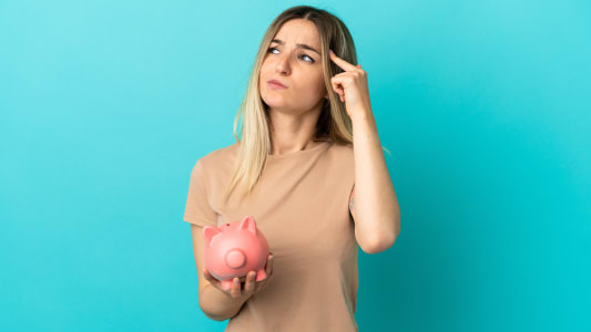 Jeune femme tenant une tirelire sur un fond bleu isol&eacute; ayant des doutes et r&eacute;fl&eacute;chissant