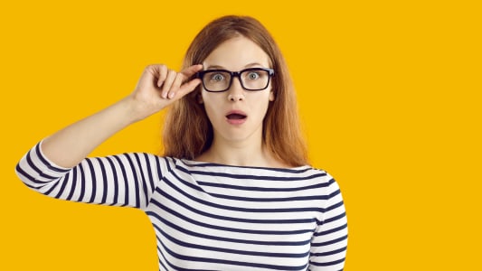 Jeune &eacute;tudiante tr&egrave;s surprise par quelque chose. Belle jeune femme nerd en haut ray&eacute; et lunettes isol&eacute;e sur fond jaune regardant la cam&eacute;ra avec une expression faciale dr&ocirc;le, choqu&eacute;e, &eacute;tonn&eacute;e