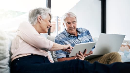 Ordinateur portable, tablette et couple de personnes &acirc;g&eacute;es sur le canap&eacute; planifiant leur fonds de retraite, leurs &eacute;conomies et leur pension. Lecture, d&eacute;tente et homme et femme &acirc;g&eacute;s avec ordinateur et technologie num&eacute;rique pour le paiement des factures &agrave; domicile.