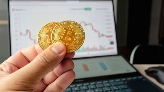 Main tenant deux bitcoins devant un ordinateur avec graphique boursier.