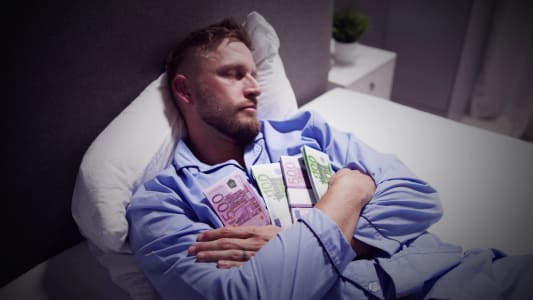 Un homme dormant dans un lit en tenant des liasses de billets dans ses bras.