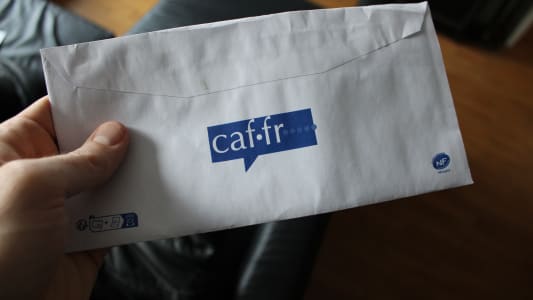 caf.fr, a hand holding an envelope with a partial view of the logo caf.fr Caisse d'Allocation Familiale on it. Translation of French: family allowance fund.