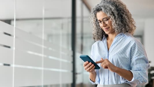 Femme d'affaires d'&acirc;ge moyen heureuse tenant un t&eacute;l&eacute;phone portable et l'utilisant au bureau. Souriant, la femme professionnelle &acirc;g&eacute;e et mature, propri&eacute;taire d'une entreprise, utilise son smartphone debout dans le couloir.