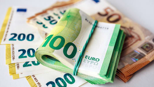 Pile de billets en euros comprenant un rouleau de billets de 100 euros sur une surface blanche avec des dénominations supplémentaires de 50 et 200 euros visibles en arrière-plan