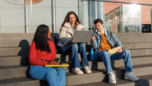 Trois étudiants assis sur des marches extérieures, utilisant un ordinateur portable et discutant.
