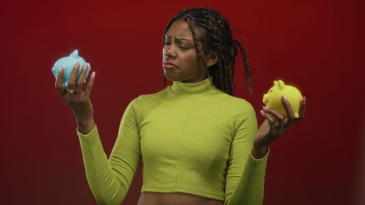 Femme tenant des tirelires bleue et jaune et fron&ccedil;ant les sourcils tout en les comparant dans un studio rouge ; incertitude financi&egrave;re.