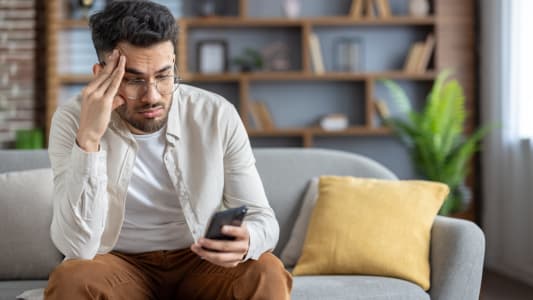 Homme frustré assis sur un canapé vérifiant son smartphone, se sentant stressé à la maison, portant des vêtements décontractés