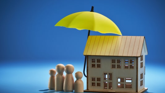 Des figurines miniatures d'une famille se tiennent &agrave; c&ocirc;t&eacute; d'une maisonsur laquelle est plac&eacute;e un parapluie pour symboliser l'assurance.