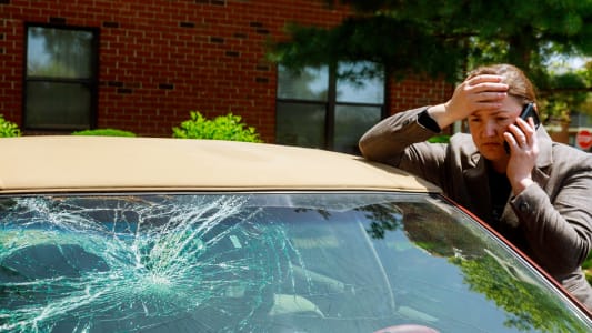 Une femme passant un appel téléphonique à côté d'une voiture dont le pare-brise est endommagé.