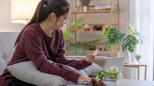 Une jeune femme de bonne humeur assise sur le canapé dans le salon et utilisant un ordinateur portable et une carte de crédit.