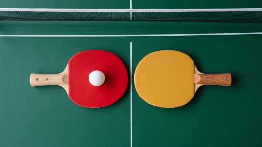 Vue de dessus de deux raquettes de ping-pong, une rouge et une jaune, avec une balle blanche sur la raquette rouge, placées sur une surface de table de tennis verte avec un filet en arrière-plan.