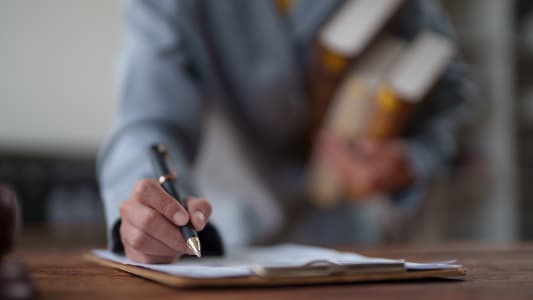 Main d'affaires tenant un stylo et signant un contrat plac&eacute; sur un presse-papiers, tout en portant &eacute;galement des livres de droit, symbolisant l'expertise juridique, l'accord et les documents officiels.