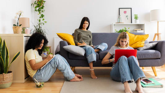 Trois &eacute;tudiantes &eacute;tudiant ensemble &agrave; la maison, assises confortablement sur le sol et le canap&eacute;, plong&eacute;es dans leurs devoirs universitaires. Concept d'amiti&eacute; f&eacute;minine et de jeunesse