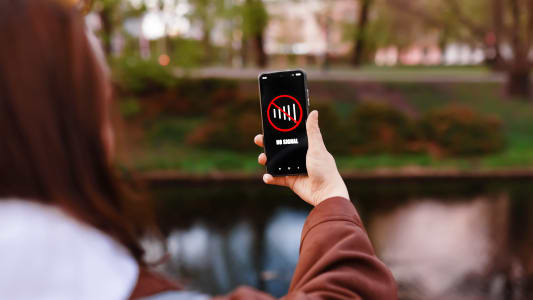 Une femme tient un smartphone affichant un message Pas de Signal tout en se tenant dans un parc pr&egrave;s de l'eau. L'image repr&eacute;sente une panne de r&eacute;seau cellulaire, une mauvaise connexion ou une perte de donn&eacute;es.