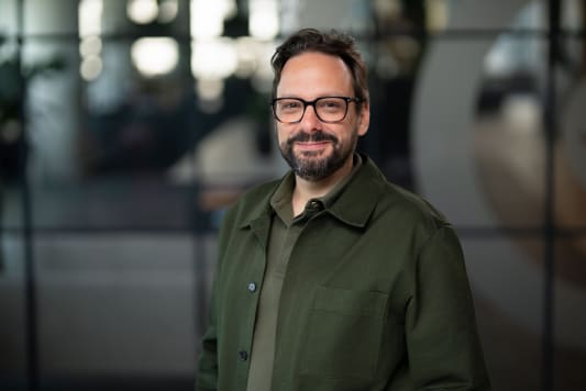 Homme souriant avec lunettes, barbe, veste verte, fond flou.